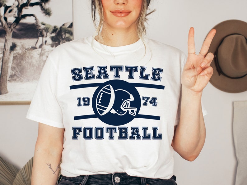 以下が含まれることがあります： 白地にネイビーブルーのグラフィックデザインが施されたTシャツ。"Seattle Football"の文字と、サークルの中にフットボールとヘルメットが描かれ、1974という数字が書かれている。