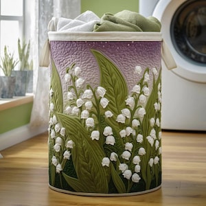 May include: A cylindrical laundry hamper with a floral design. The hamper features a textured surface with white lily of the valley flowers and green leaves against a purple and green background. White and green towels are visible inside.