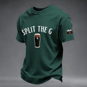 Pode incluir: T-shirt verde escuro com o texto "SPLIT THE G" acima de um desenho de um pint de stout. A letra "G" está no centro do pint. Duas canecas de cerveja castanhas são bordadas na manga.