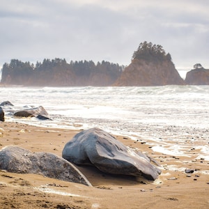 May include: A rocky beach with large grey boulders in the foreground, a sandy shore, and a forested island in the distance. The ocean waves are crashing on the shore.