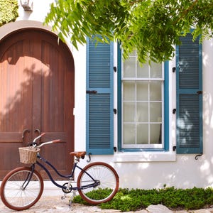 May include: A navy blue bicycle with a wicker basket and brown tires is parked in front of a white building with teal shutters and a wooden door. Green foliage and a tree branch frame the scene.