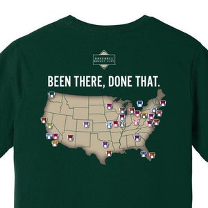 May include: A dark green t-shirt displaying the text "Been There, Done That" above a map of the United States. The map is adorned with small, colourful icons. The shirt also features the text "Baseball Bucket List".