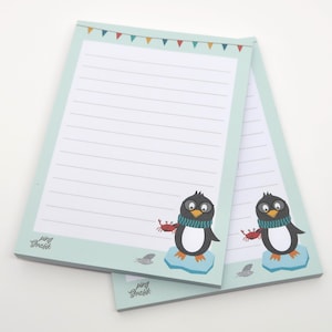 May include: Two lined notepads with a light teal border and a cartoon penguin illustration. The penguin wears a blue and teal scarf and holds a red crab. The top of the notepad has a banner of colorful flags. The text "ping Grafik" is at the bottom.