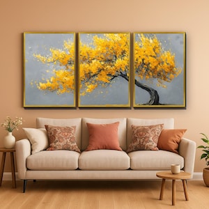 May include: A triptych wall art set featuring a single tree with vibrant yellow leaves against a solid grey background, framed in thin gold. The three rectangular canvases are arranged horizontally above a beige sofa with decorative cushions.