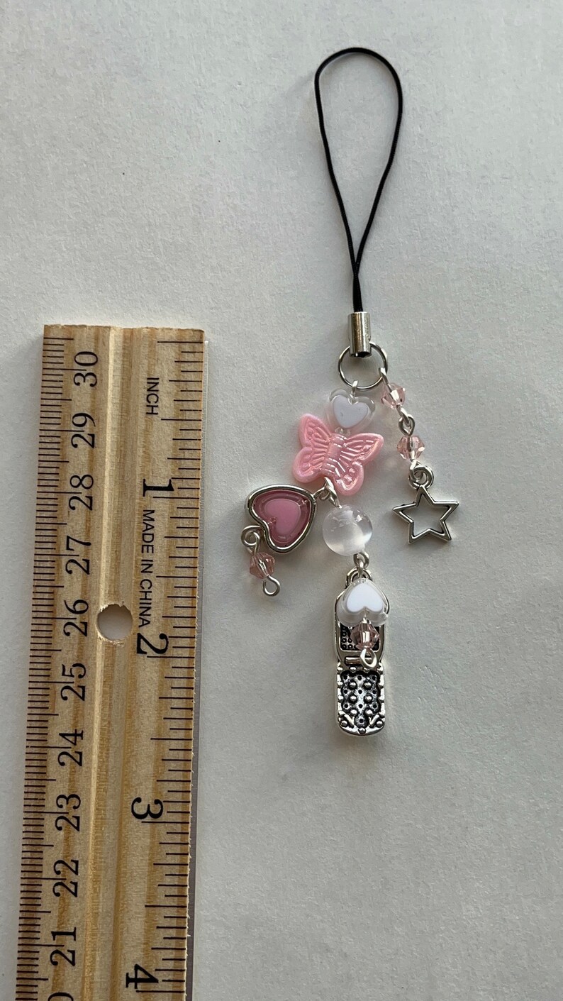 May include: A pink and white butterfly charm with a silver heart, star, and phone charm. The charms are attached to a black string with a silver clasp.
