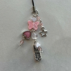May include: A pink and white cell phone charm with a butterfly, hearts, a star, and beads. The charm is attached to a black string.