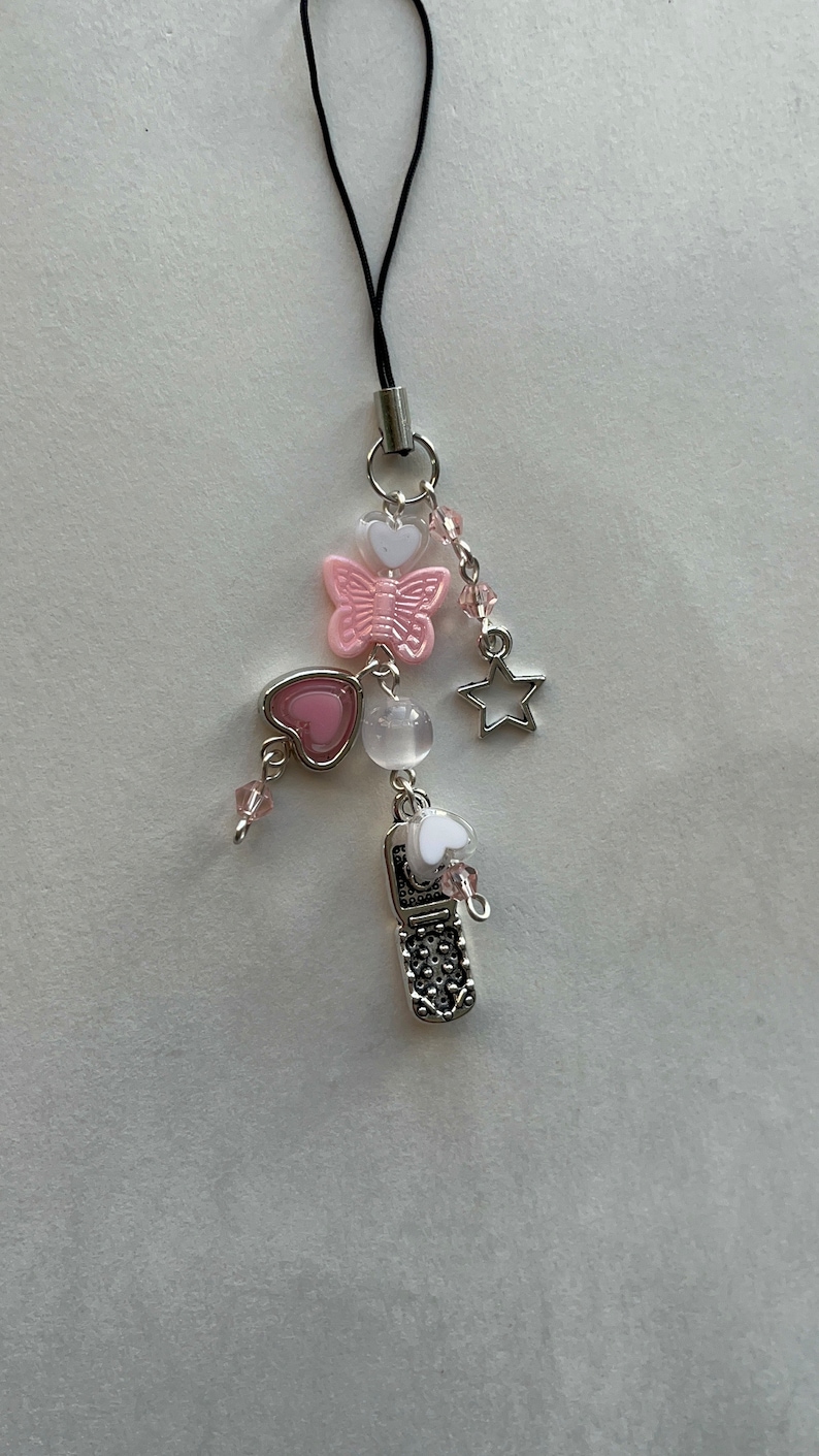 May include: A pink and white cell phone charm with a butterfly, hearts, a star, and beads. The charm is attached to a black string.