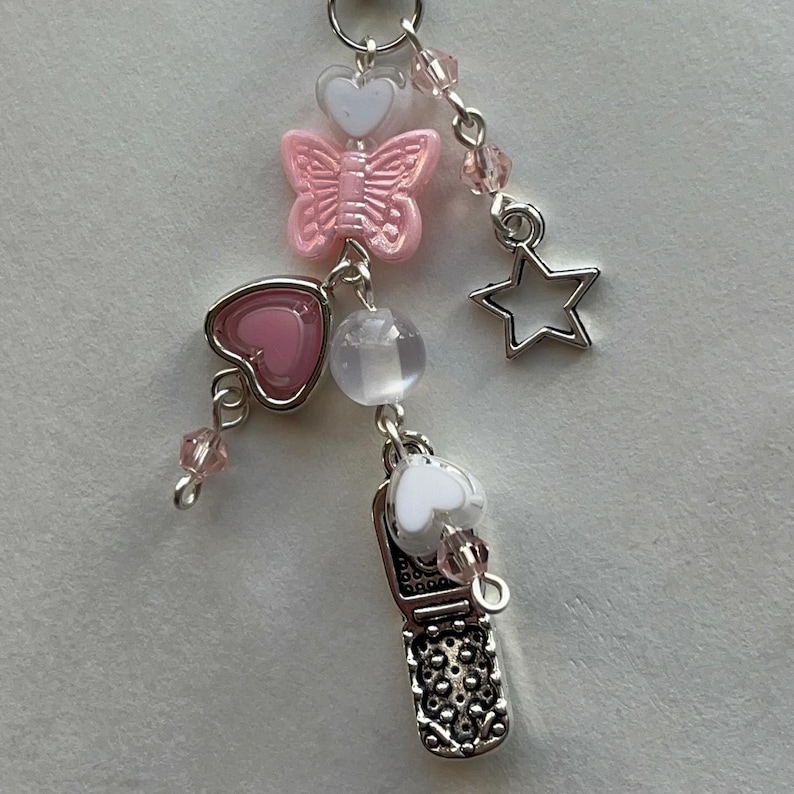 May include: A silver charm with a pink butterfly, pink heart, white heart, clear bead, silver star, and a silver flip phone charm.