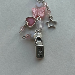 May include: A silver charm with a pink butterfly, two pink hearts, a white heart, a white bead, a silver star, and a silver charm shaped like a lipstick tube. The charm is attached to a black phone strap.