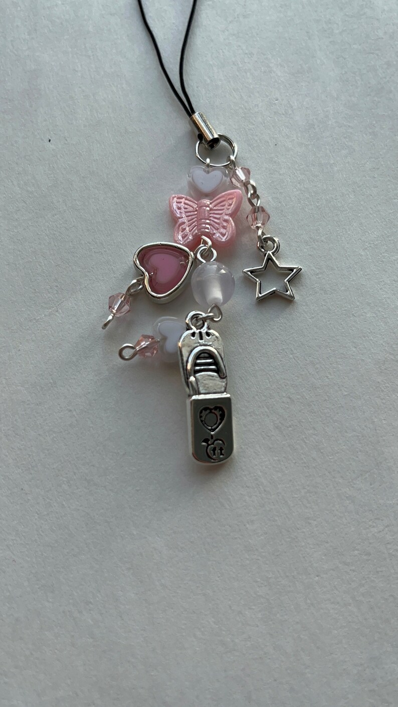 May include: A silver charm with a pink butterfly, two pink hearts, a white heart, a white bead, a silver star, and a silver charm shaped like a lipstick tube. The charm is attached to a black phone strap.