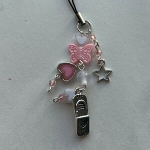 May include: A pink and white charm with a butterfly, hearts, and a star. The charm is attached to a black string.