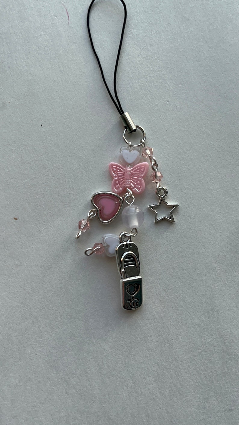 May include: A pink and white charm with a butterfly, hearts, and a star. The charm is attached to a black string.