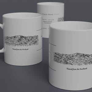 May include: White ceramic mugs with black line drawings of mountain ranges. One mug is labeled "Viewed from the Northeast," another "Viewed from the Southwest," and a third mug has text that reads "Pikes Peak, Front Range, Colorado."