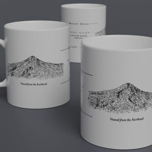 May include: Two white ceramic mugs featuring black line drawings of Mount Hood, a mountain in Oregon. The mugs depict the mountain from two different viewpoints, "Viewed from the Southwest" and "Viewed from the Northeast".
