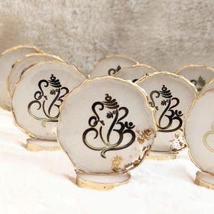 Op de afbeelding: Een verzameling witte hars onderzetters met gouden randen en voetjes. Elke onderzetter heeft een zwarte illustratie van de hindoeïstische godheid Ganesha. Sommige onderzetters hebben bladgoud accenten, wat een elegante en spirituele uitstraling creëert.