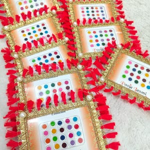 Puede incluir: Surtido de pegatinas bindi de colores en una variedad de colores y diseños. Las pegatinas están empaquetadas en pequeños marcos cuadrados con ribetes rojos y dorados y el texto "Pooja Laxmi" impreso en la parte inferior.