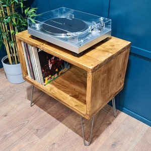May include: A rectangular wooden record player stand with a natural brown finish and four thin silver metal hairpin legs. The top surface holds a silver turntable with a clear plastic dust cover, while the open shelf stores vinyl records.