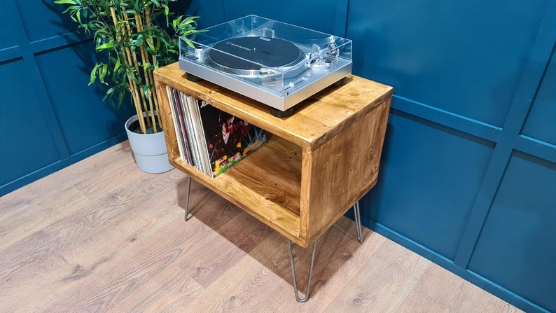 May include: A rectangular wooden record player stand with a natural brown finish and four thin silver metal hairpin legs. The top surface holds a silver turntable with a clear plastic dust cover, while the open shelf stores vinyl records.