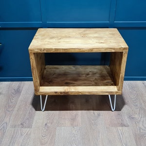 May include: A rectangular wooden side table with a natural brown wood grain finish, featuring an open storage shelf and four thin white metal hairpin legs. The table has a smooth top surface and a matching lower shelf for storage.