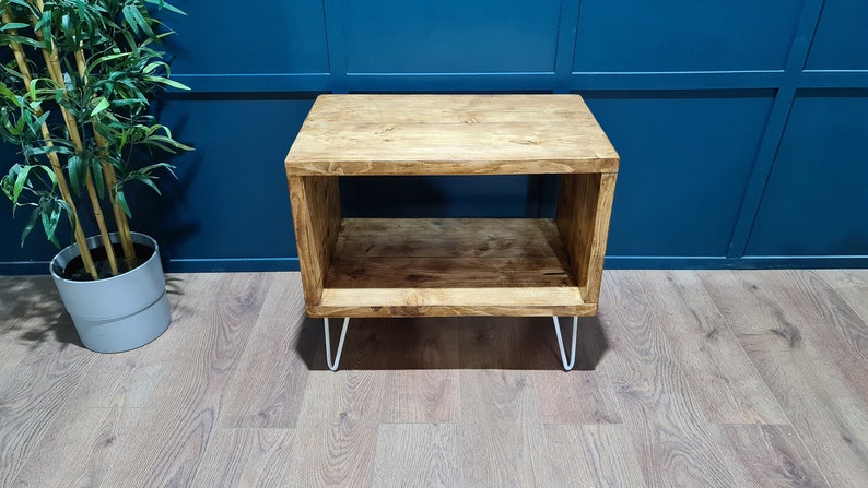 May include: A rectangular wooden side table with a natural brown wood grain finish, featuring an open storage shelf and four thin white metal hairpin legs. The table has a smooth top surface and a matching lower shelf for storage.
