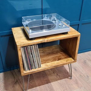 May include: A silver Audio-Technica turntable with a clear dust cover sits atop a rectangular wooden storage unit with four thin metal hairpin legs. The unit features an open shelf containing a collection of vinyl records stored vertically.