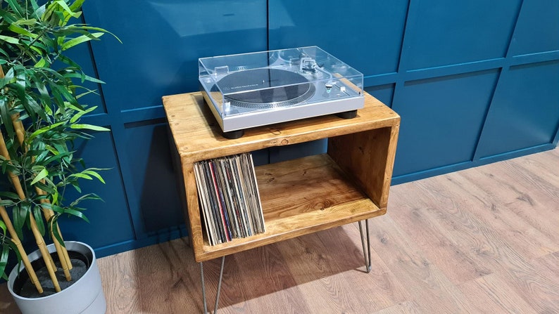 May include: A silver Audio-Technica turntable with a clear dust cover sits atop a rectangular wooden storage unit with four thin metal hairpin legs. The unit features an open shelf containing a collection of vinyl records stored vertically.