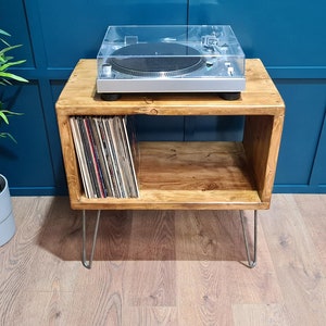 May include: A rectangular wooden record player stand with a natural brown finish and four thin metal hairpin legs. The top surface holds a silver turntable with a clear plastic cover, while the open storage shelf contains a collection of vinyl records.