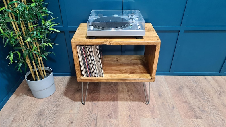 May include: A rectangular wooden record player stand with a natural brown finish and four thin metal hairpin legs. The top surface holds a silver turntable with a clear plastic cover, while the open storage shelf contains a collection of vinyl records.