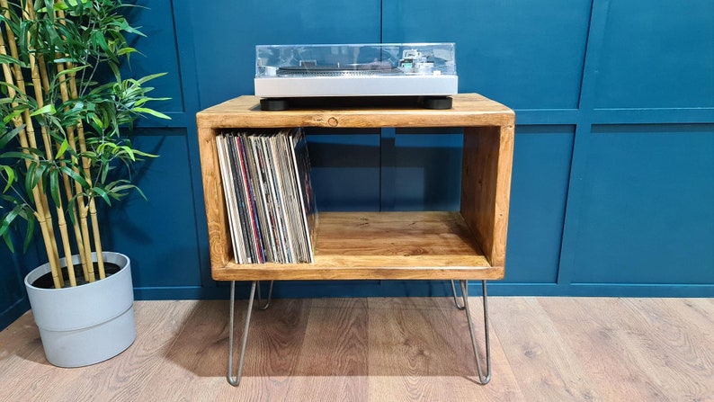 May include: A rectangular wooden storage unit with a natural grain finish, supported by four thin silver metal hairpin legs. The unit features an open shelf containing a collection of vinyl records, with a silver turntable placed on the top surface.