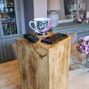 May include: A square wooden side table with a natural finish. A black remote, a smartphone, and a mug with a black and pink design are on top. The table is made of light-coloured wood with visible grain. The table is approximately 40 cm tall.