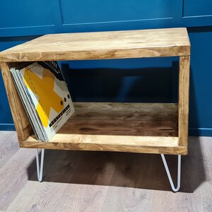 May include: A rectangular wooden storage unit with a natural brown wood grain finish, featuring an open shelf design and four thin white metal hairpin legs. A collection of vinyl records with a yellow graphic cover is stored inside the unit.