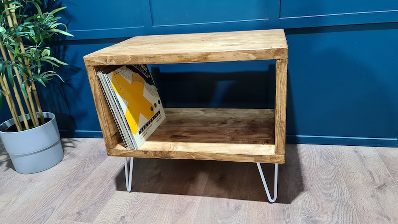 May include: A rectangular wooden storage unit with a natural brown wood grain finish, featuring an open shelf design and four thin white metal hairpin legs. A collection of vinyl records with a yellow graphic cover is stored inside the unit.