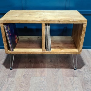 May include: Wooden record storage unit with two compartments, each holding vinyl records. The unit is made of light-coloured wood and has metal hairpin legs. The background is a dark blue wall and a light wood floor.