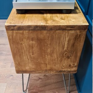 May include: A rectangular wooden side table with a natural grain finish, supported by four thin metal hairpin legs. A silver turntable with a clear plastic dust cover is positioned on the flat top surface of the wooden table.