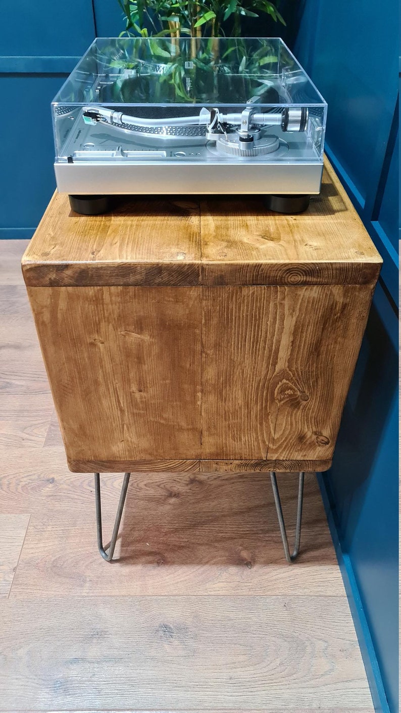 May include: A rectangular wooden side table with a natural grain finish, supported by four thin metal hairpin legs. A silver turntable with a clear plastic dust cover is positioned on the flat top surface of the wooden table.
