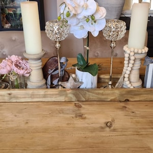 May include: A wooden tray arrangement featuring candles, decorative orbs, and artificial flowers. The rectangular tray sits on a wooden table. The display includes various textures and colours, creating a rustic and elegant centrepiece for a home.