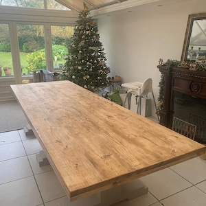 May include: A large rectangular dining table with a natural light brown wood grain finish and a thick tabletop. The table is supported by two sturdy, light grey pedestal bases that sit directly on the white tiled floor of the room.