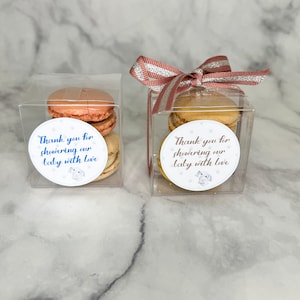 Peut inclure: Deux boîtes transparentes avec des nœuds en ruban rose et or contiennent des piles de macarons. Chaque boîte a un autocollant rond avec le texte "Merci d'avoir gâté notre bébé avec amour" et une petite illustration d'un chien.
