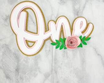 Könnte beinhalten: Ein rosa und goldener "One"-Kuchentopper mit einer rosa Papierblume und grünen Blättern.