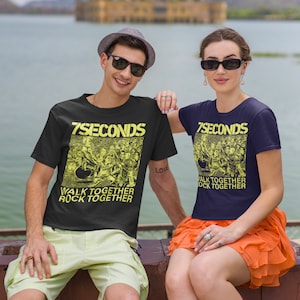 May include: A couple wearing matching black t-shirts with a yellow graphic of a band called "7 Seconds" and the text "Walk Together Rock Together". The man is wearing a brown hat and sunglasses. The woman is wearing a bright orange skirt.