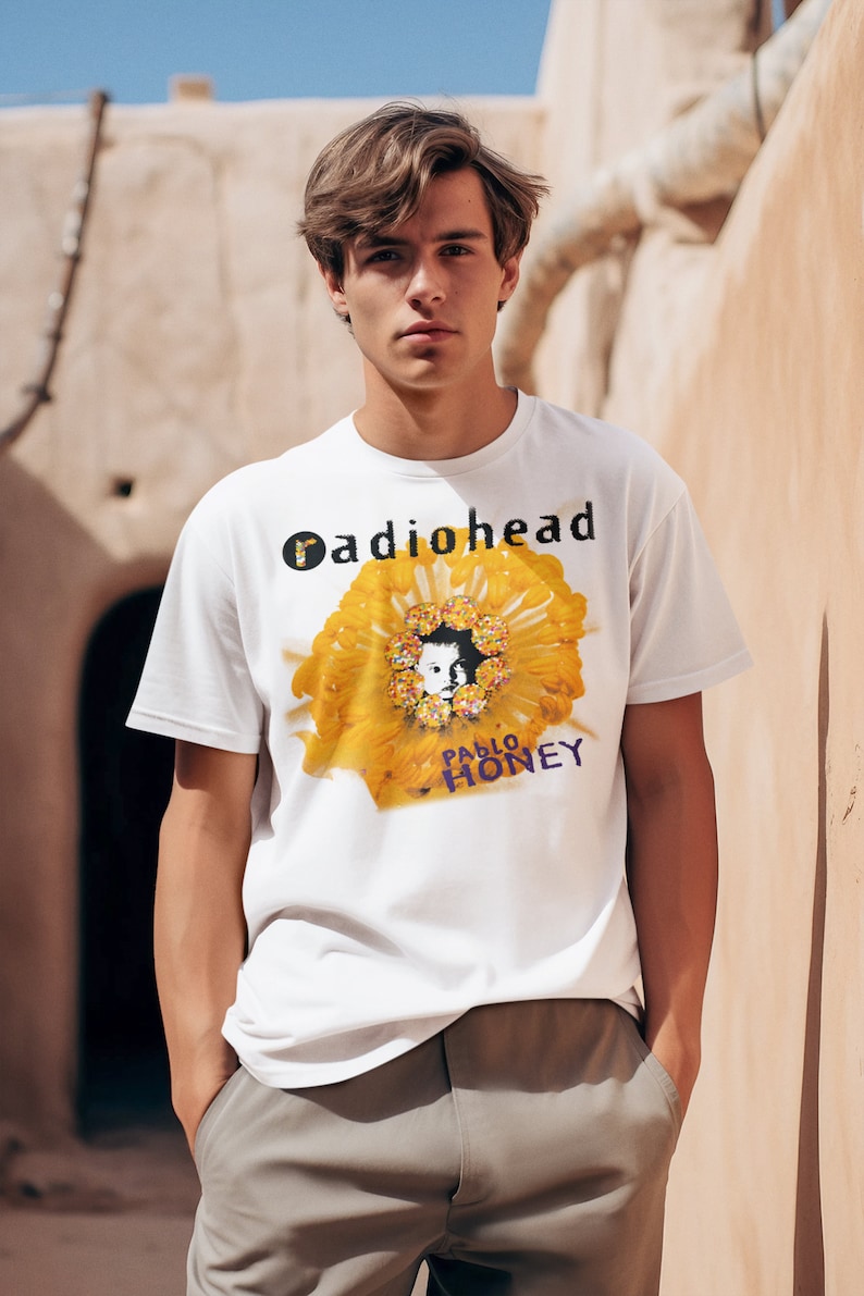 Pu&ograve; includere: Una maglietta bianca con una grafica di un fiore giallo e il testo "Radiohead" e "Pablo Honey" stampato sul davanti.