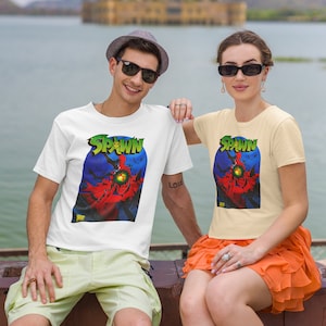 May include: Two people wearing white and cream t-shirts with a graphic print of Spawn, a comic book character. The graphic is a close-up of Spawn's face with a red cape and a green eye. The text "Spawn" is printed in black above the graphic. The people are sitting on a stone wall with a lake and a building in the background.