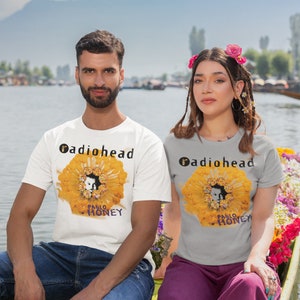 Pu&ograve; includere: Due persone indossano magliette con la copertina dell'album dei Radiohead "Pablo Honey" stampata su di esse. La copertina dell'album presenta un fiore giallo con il viso di una persona al centro. Le persone sono sedute su una barca su un lago.