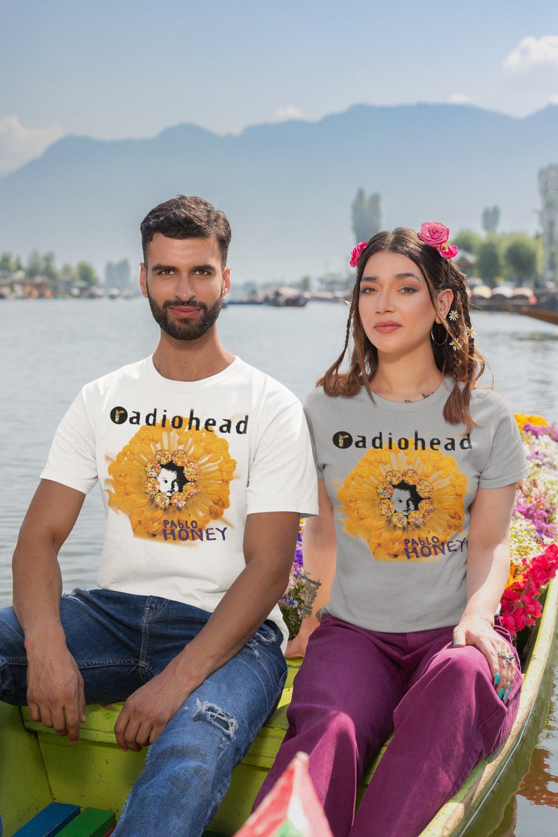 Pu&ograve; includere: Due persone indossano magliette con la copertina dell'album dei Radiohead "Pablo Honey" stampata su di esse. La copertina dell'album presenta un fiore giallo con il viso di una persona al centro. Le persone sono sedute su una barca su un lago.