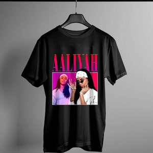 May include: Black t-shirt with a pink and white graphic of singer Aaliyah. The graphic features a portrait of Aaliyah wearing a white bandana and sunglasses.