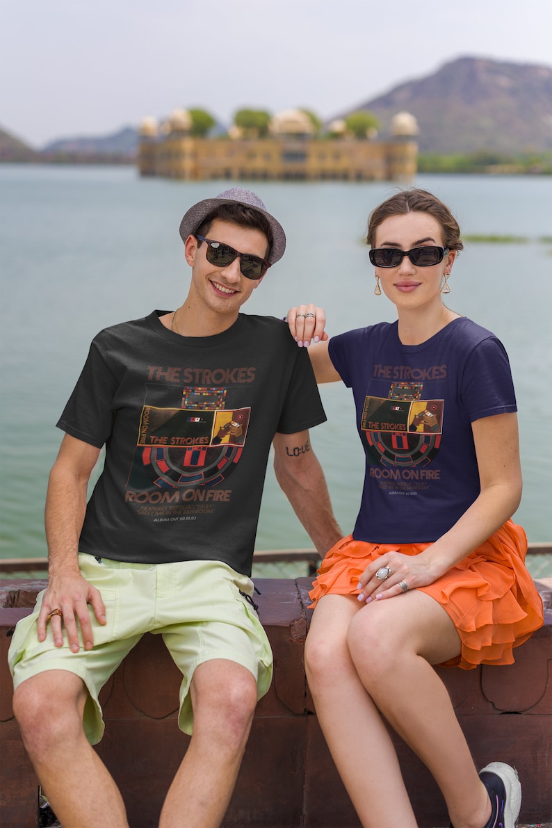 May include: Two people wearing black t-shirts with a graphic of The Strokes band logo and the text "Room On Fire". The man is wearing a light green pair of shorts and a hat. The woman is wearing a dark blue t-shirt and an orange skirt.