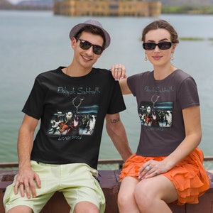 May include: Two people wearing black t-shirts with a Black Sabbath "Live Evil" album cover design. The man is wearing a light green pair of shorts and a brown hat. The woman is wearing a bright orange skirt.