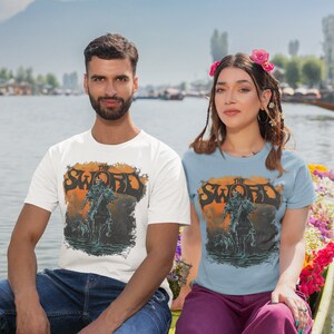 May include: Two people wearing t-shirts with a graphic of a person riding a horse and the text "The Sword". The person on the left is wearing a white t-shirt and the person on the right is wearing a light blue t-shirt. They are sitting in a boat on a lake.