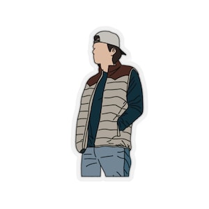 Op de afbeelding: Een man draagt een beige en bruine puffer vest met een blauw longsleeve shirt eronder. Hij draagt een witte baseball cap en een blauwe jeans.