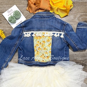 May include: A denim jacket with a floral patch on the back and white fringe trim. The jacket is paired with a white tulle skirt.